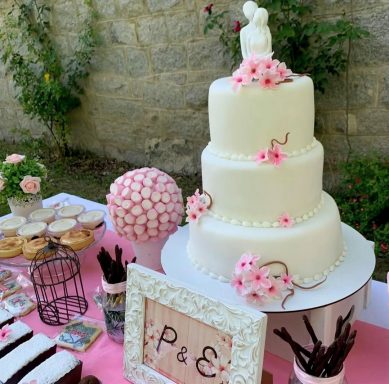 Tarta blanca de tres pisos decorada con flores y figuras de novios, acompañada de dulces.