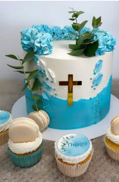 Tarta blanca decorada con flores azules, hojas y un crucifijo, acompañada de cupcakes.