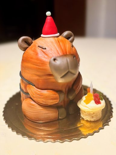 Tarta en forma de un capibara con gorro navideño y cupcake al lado.