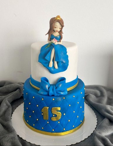 Tarta de dos pisos azul, con figura de una princesa y decoraciones doradas.